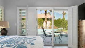 A bedroom with a bed, a nightstand, a lamp, a sliding glass door to a patio with a pool, chairs, and a wooden dresser.