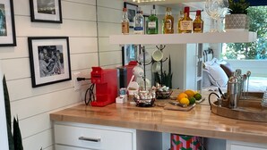 Une cuisine moderne avec des meubles blancs, un plan de travail en bois et un mini-réfrigérateur encastré. On peut voir des tableaux encadrés au mur, une machine à café, ainsi qu’une variété de bouteilles et de verres sur le plan de travail.