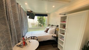 Une chambre d’hôtel de taille modeste, comprenant un lit, une petite table avec une bouteille et des verres, une chaise, un bureau avec une lampe et une bibliothèque.