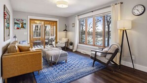 A cozy living room with a sofa, ottoman, and a chair. There is a large window with curtains, a clock on the wall, and a lamp. - Gratitude Up North On Round Lake (Hayward)