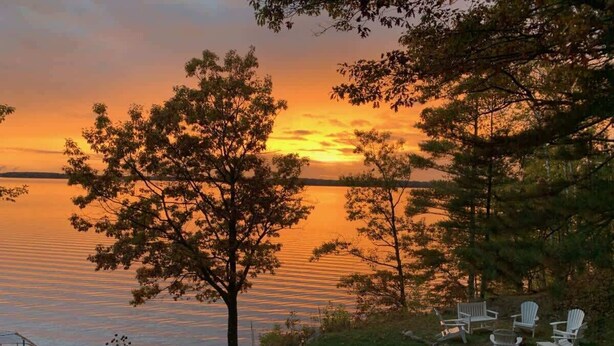 A lakeside view with a sunset, trees, and white Adirondack chairs. - Gratitude Up North On Round Lake (Hayward)