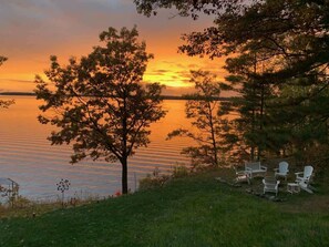 A lakeside view with a sunset, trees, and white Adirondack chairs. - Gratitude Up North On Round Lake (Hayward)