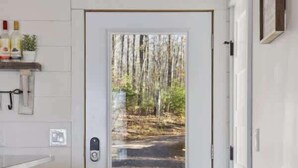 A modern interior with a white door leading to a forest view, a small shelf with bottles, and a red stool. - Gratitude Up North On Round Lake (Hayward)