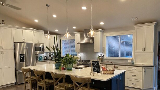 Una cocina moderna con armarios blancos, un refrigerador de acero inoxidable, una isla grande con taburetes, luces colgantes y un letrero de pizarra.