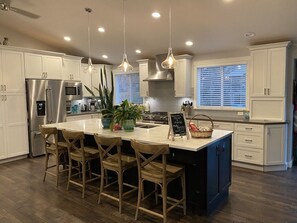 Una cocina moderna con armarios blancos, un refrigerador de acero inoxidable, una isla grande con taburetes, luces colgantes y un letrero de pizarra.