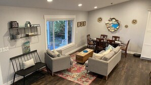 Una sala de estar con un conjunto de sofá, una mesa de centro y una mesa de comedor con sillas. Hay una ventana que ofrece vistas al exterior.