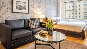 A modern hotel room with a black leather sofa, a glass coffee table with books and a vase of flowers, a bed with a white comforter, and a large window with a view of a building.
