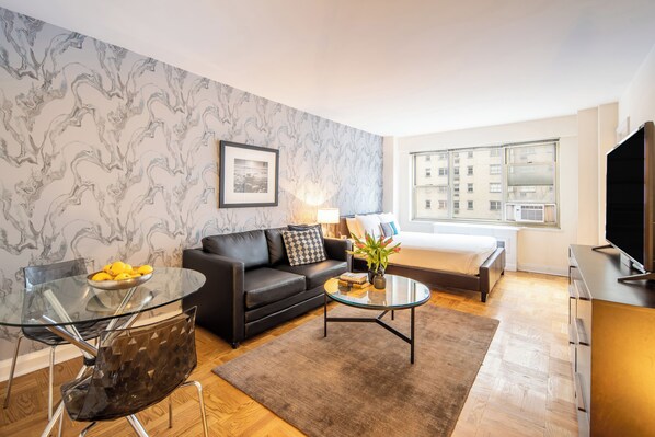 A modern living room with a glass coffee table, a black leather sofa, a glass dining table with chairs, and a large window offering a city view.