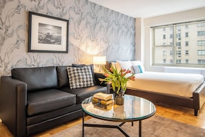 A modern hotel room with a black leather sofa, a glass coffee table with books and a vase of flowers, a bed with a white comforter, and a large window with a view of a building.