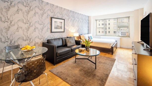 A modern living room with a glass coffee table, a black leather sofa, a glass dining table with chairs, and a large window offering a city view.