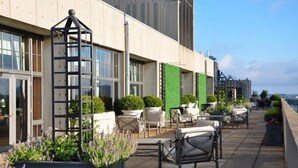 Un área de estar al aire libre con sillas, macetas y una estructura metálica, junto a la fachada de un edificio con ventanas y un muro verde.