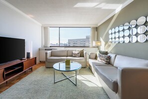 A modern living room with a flat-screen TV, two sofas, a glass coffee table, and a city view through large windows.