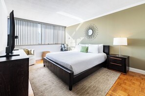 A modern hotel room with a large bed, a flat-screen TV, a bedside table with a lamp, and a view of the outside through the window. - Beautiful Two Bedroom near Prudential Center (Boston)