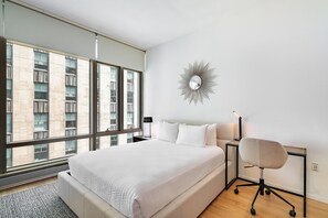 A modern bedroom with a large bed, a desk, a chair, and a view of buildings through large windows. - Bright Escape | Fifth Ave | W/D & Gym (New York)