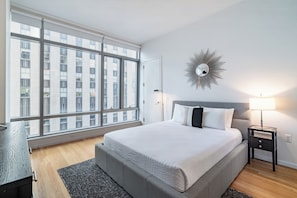 Ein modernes Hotelzimmer mit großem Fenster, einem Bett mit grauer Bettwäsche, einem Nachttisch mit Lampe und einem an der Wand befestigten Spiegel.