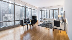 A modern living room with large windows, a dining area, and a sofa. - Luxury Stay | Near Rockefeller | City Views & W/D (New York)