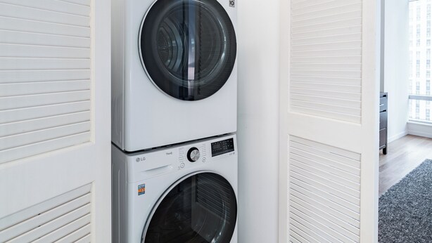 A laundry room with two stacked LG washing machines, white walls, wooden flooring, and a white door. - Luxury Stay | Near Rockefeller | City Views & W/D (New York)