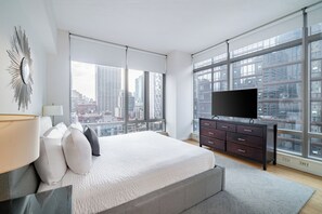 A modern bedroom with a large bed, a TV, and a city view through floor-to-ceiling windows. - Luxury Stay | Near Rockefeller | City Views & W/D (New York)