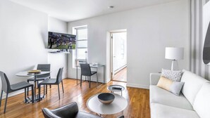 A modern living room with a flat-screen TV, a dining area with a glass table and chairs, a sofa, and a lamp.