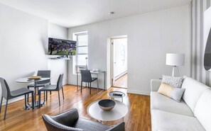 A modern living room with a flat-screen TV, a dining area with a glass table and chairs, a sofa, and a lamp.