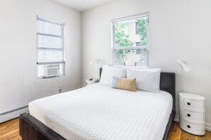 A bedroom with a bed, two bedside tables, a window with blinds, and a wall-mounted lamp.