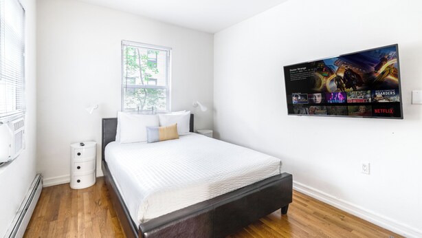 A bedroom with a bed, a window, a wall-mounted TV displaying various shows, and a white wall.