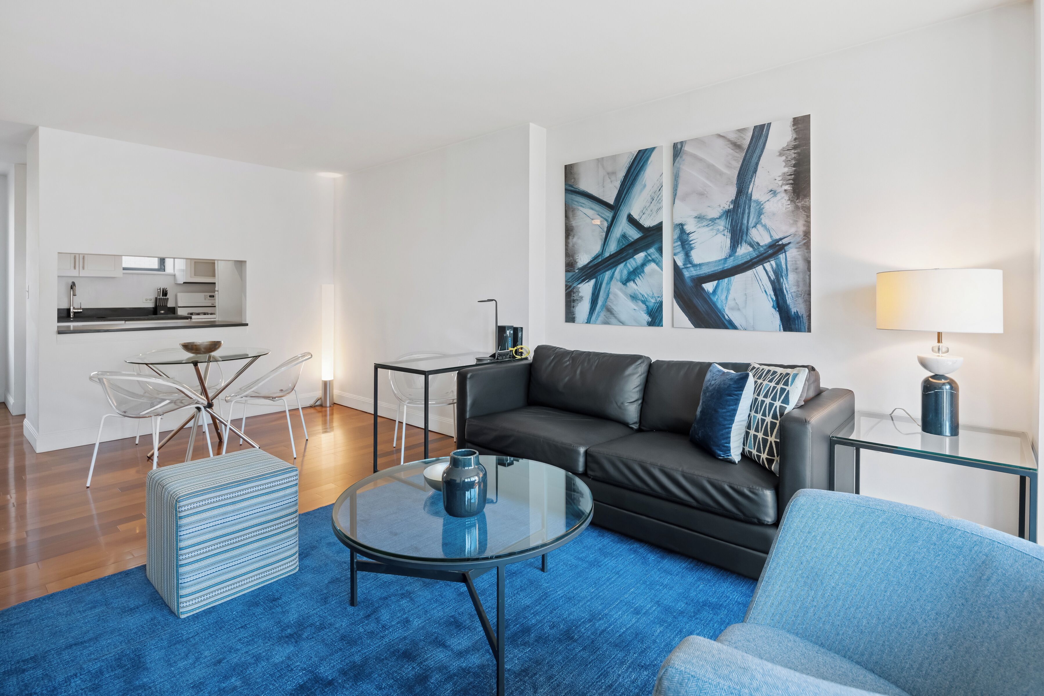 A modern living room with a black leather sofa, a glass coffee table, a blue patterned ottoman, and a blue armchair. There is a kitchen area with a dining table and chairs visible in the background.