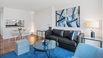 A modern living room with a black leather sofa, a glass coffee table, a blue patterned ottoman, and a blue armchair. There is a kitchen area with a dining table and chairs visible in the background.