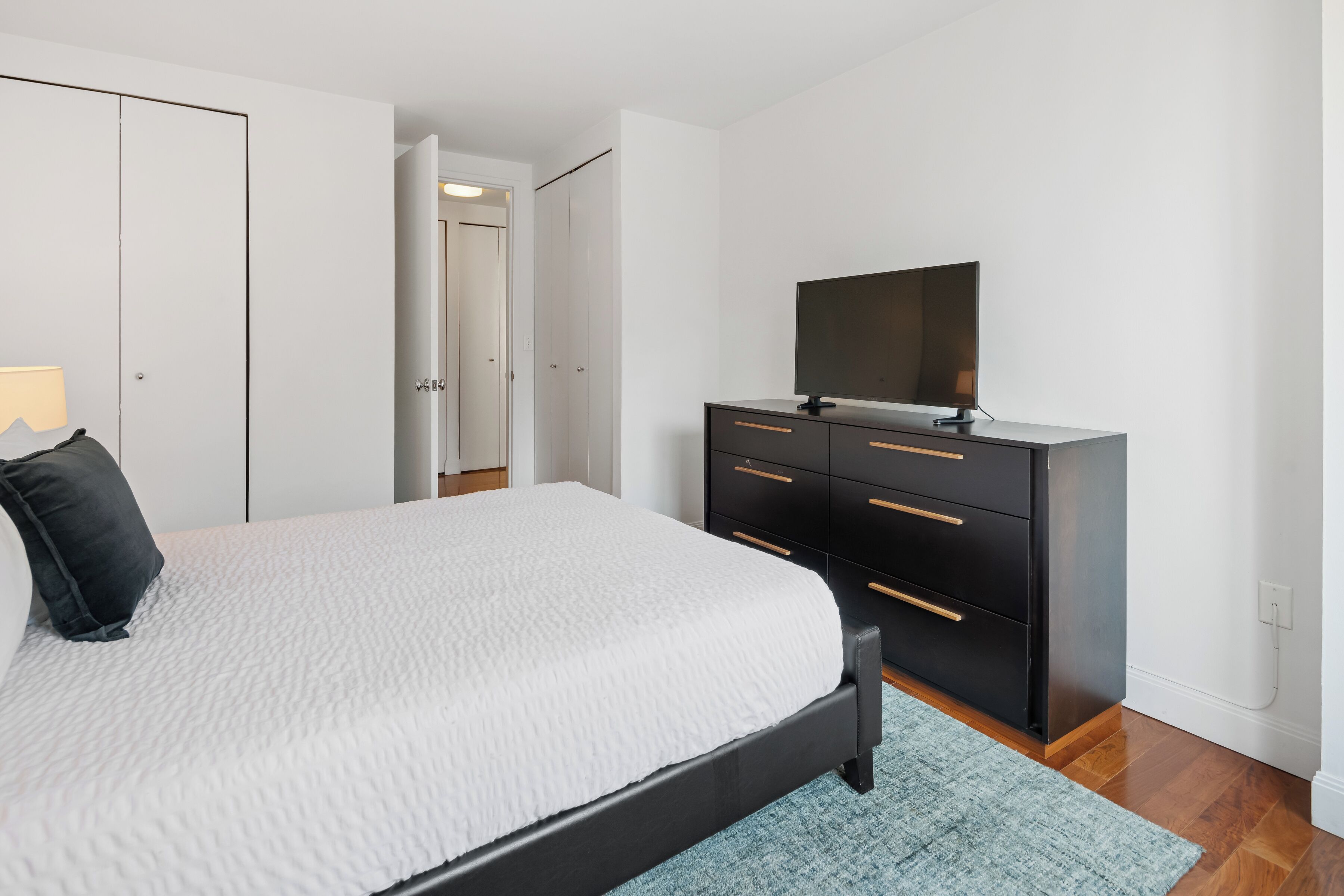 A bedroom with a bed, a black dresser, a television, and a door leading to another room.