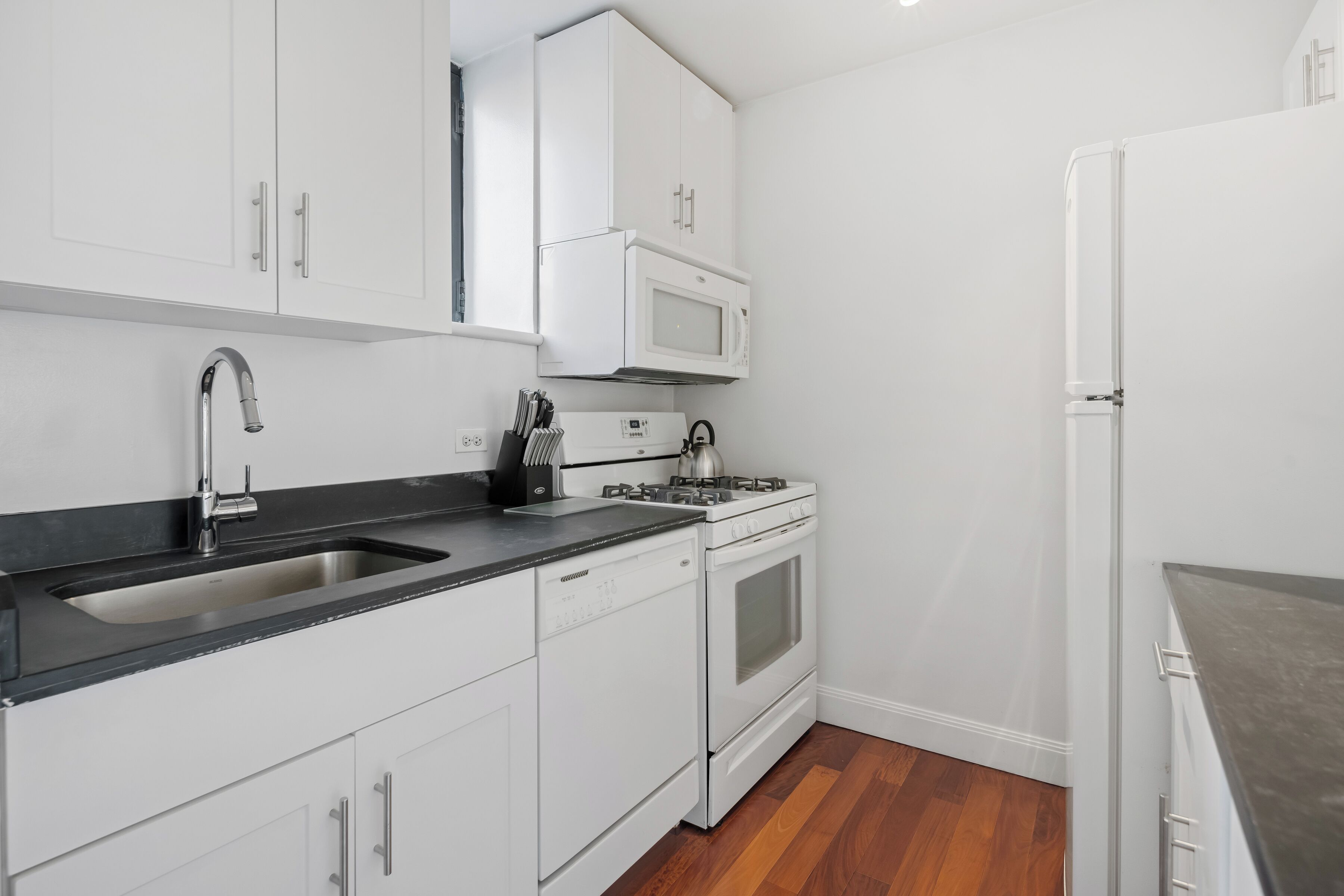 A modern kitchen with white cabinets, a black countertop, a stainless steel sink, a microwave, a stove, and a refrigerator.