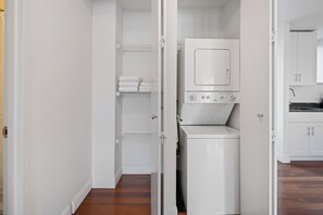 A laundry room with a stacked washer and dryer, a shelf with folded towels, and a white door leading to a kitchen area. - Contemporary Gramercy Stay | Balcony, Rooftop Deck (New York)