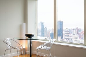 A modern room with a glass table and two transparent chairs, a white vase, and a cityscape view through large windows.
