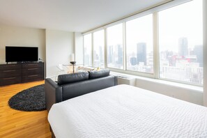 A modern hotel room with a large bed, a flat-screen TV, a dark-colored sofa, a round rug, and a city view through large windows.
