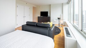 Et moderne hotelværelse med en stor seng, en sort lædersofa, et lille glasbord, et fjernsyn og udsigt over byen.