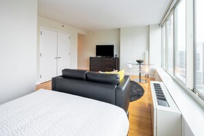 A modern hotel room with a large bed, a black leather sofa, a small glass table, a TV, and a city view.