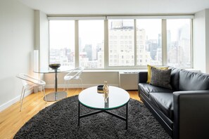 A modern living room with a glass coffee table, a black leather sofa, a black rug, and a city view through large windows.