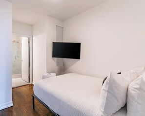 Un dormitorio con cama, televisor montado en la pared, puerta que da al baño y una mesita de noche.