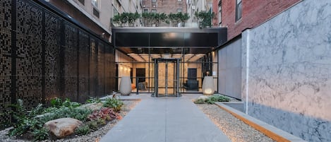A modern building entrance with a glass door, a pathway lined with pebbles, and a brick wall on one side.