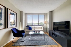 Un salon moderne avec un canapé, un fauteuil bleu, une table basse, un téléviseur et des œuvres d’art encadrées accrochées au mur.