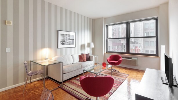 Un salon moderne avec un canapé blanc, une table basse en verre et des fauteuils rouges. Une grande fenêtre offre une vue sur la ville, un tableau encadré orne le mur et une lampe est posée sur la table.