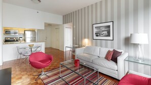 Un salon moderne avec un canapé blanc, un fauteuil rouge et une table basse en verre. On aperçoit, au fond, une cuisine équipée d’appareils électroménagers en acier inoxydable.