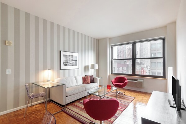 A modern living room with a white sofa, glass coffee table, and red chairs. There is a large window with a city view, a framed picture on the wall, and a lamp on the table.
