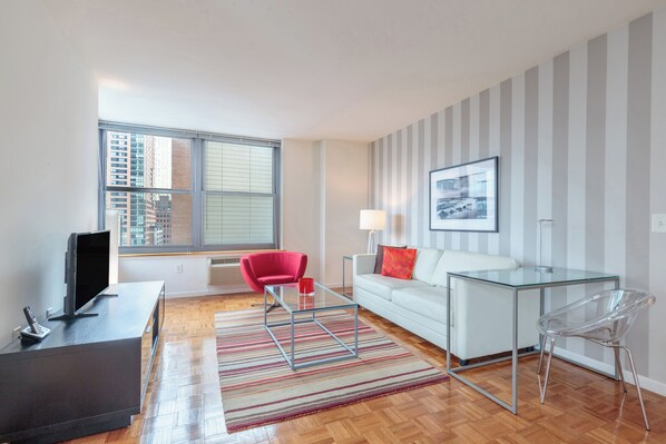 A modern living room with a sofa, a glass coffee table, a red chair, a TV on a stand, and a window with a city view.