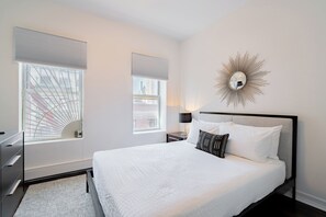 A modern bedroom with a large bed, two bedside tables, a wall-mounted mirror, and a view of the city through the windows. - Charming Two Bedroom Conveniently Located on UES (New York)
