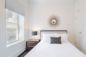 Ein Schlafzimmer mit einem Bett, einem Nachttisch, einer Lampe, einem Wandspiegel und einem Fenster mit Jalousien.