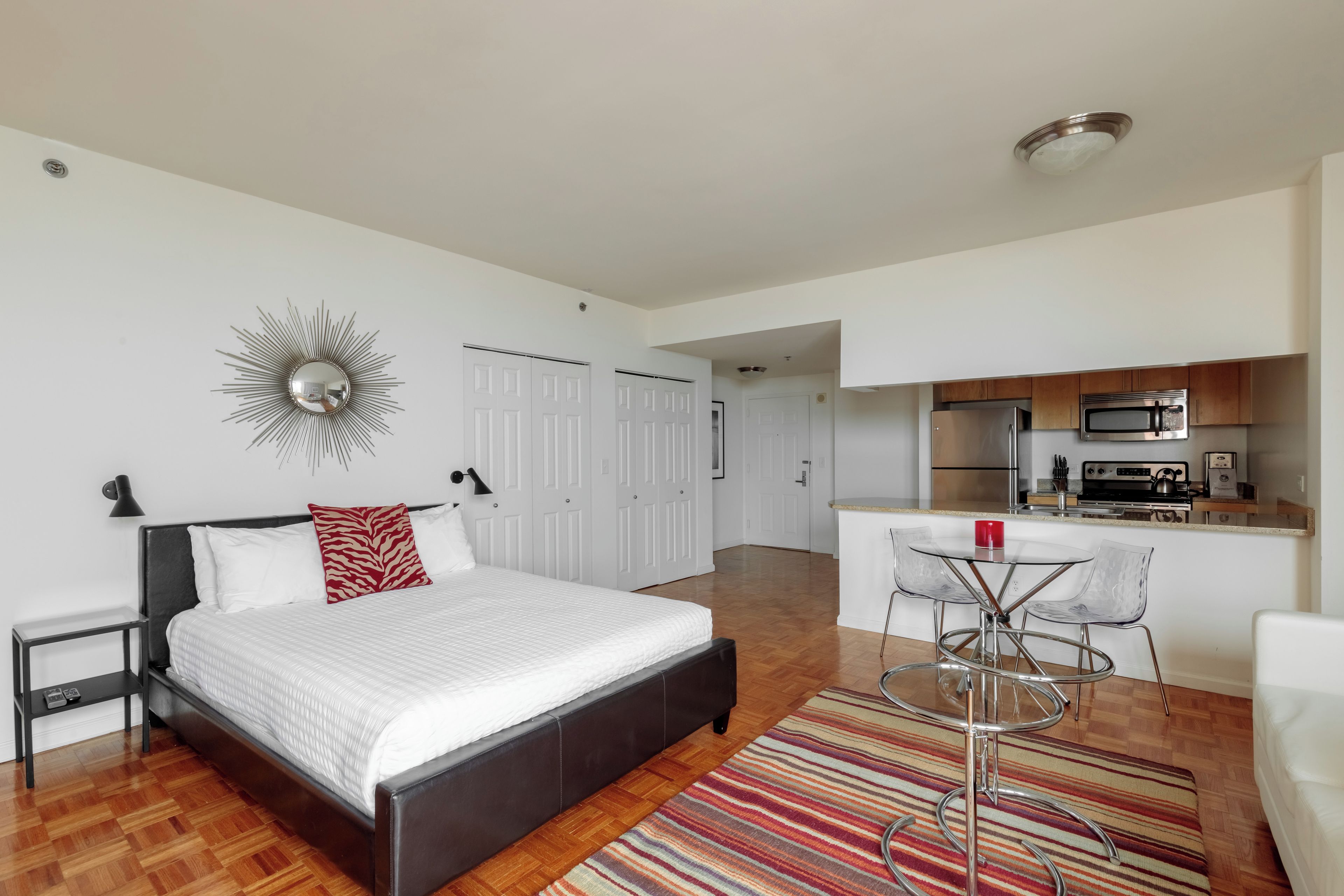 A modern bedroom with a large bed, a bedside table, a red and white pillow, a wall-mounted mirror, and a kitchen area with a dining table and chairs.