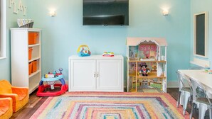Una sala de juegos infantil con un televisor montado en la pared, una alberca para bebés, una casita de muñecas y una alfombra colorida.