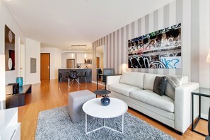 Ein modernes Wohnzimmer mit einem weißen Sofa, einem grauen Hocker und einem runden Couchtisch. Im Hintergrund ist eine Küche erkennbar, über der Küchenzeile hängt ein Wandbild mit Streifenmuster.