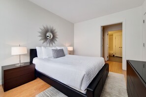 Ein modernes Schlafzimmer mit einem großen Bett, dunklen Nachttischen, einem an der Wand befestigten Spiegel und einer Tür zu einem anderen Raum.