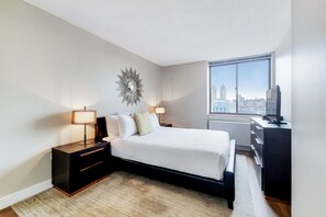 Ein modernes Schlafzimmer mit einem großen Bett, Nachttischlampen, einer Kommode und Blick auf eine Stadtlandschaft durch ein großes Fenster.